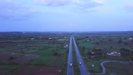 Aerial-drone-shot-of-goods-carrier-trucks-on-a-Highway-road-through-farms-in-rural-Gwalior-of-India-during-Twilight-time