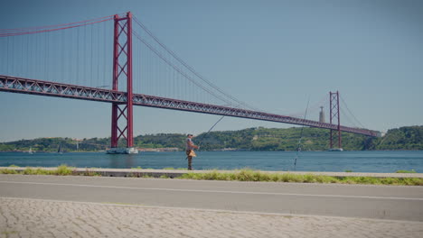 Man-Fishing-In-Tagus-River-With-25-April-Bridge-In-The-Background-May-2023