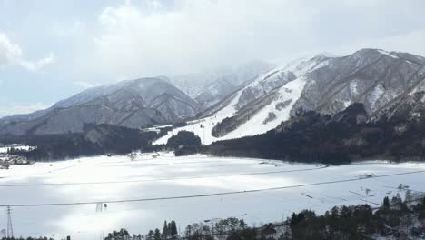 The-drone-gracefully-captures-a-mesmerizing-aerial-view-of-Hakuba-valley-and-the-vast-snow-field-in-Japan-during-winter,-showcasing-a-captivating-landscape-adorned-with-pristine-snow-and-lush-forests