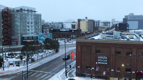 Día-De-Invierno-Helado-En-El-Centro-De-Denver-Misión-De-Rescate-Estancia-Para-Personas-Sin-Hogar-Pájaro-Gris-Nevando-Tráfico-Pasando-Edificio-Jesús-Salva-Drone-Cinematográfico-Antena-Panorámica-Círculo-A-La-Derecha