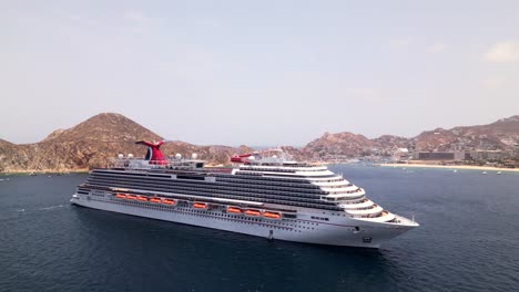 An-aerial-drone-circles-the-Carnival-Panorama-cruise-ship-anchored-in-the-bay-with-the-city-of-Cabo-San-Lucas,-Mexico-in-the-background