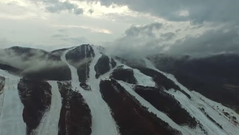 Slow-motion-drone-view-slowly-zooming-in-on-partially-snow-covered-mountains,-overcast-sky-and-fluffy-clouds
