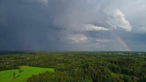 Drone-view-flying-over-a-gorgeous-countryside-of-lush-trees-and-grass,-moving-towards-storm-clouds-and-a-rainbow