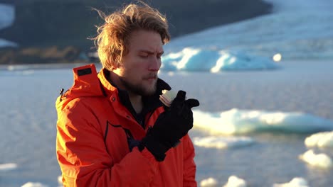 Blonde-Man-in-Orange-Coat-Eats-a-Sandwich-at-Jökulsárlón-Icebergs-Iceland-Snowy-Landscape