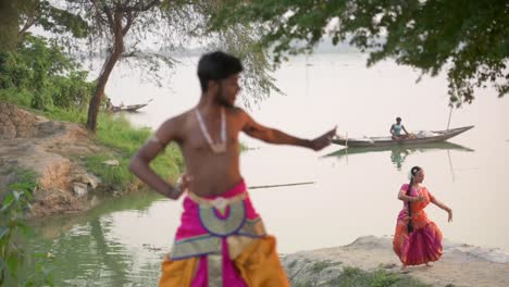 Bailarines-Indios-Masculinos-Y-Femeninos-Bailando-Danza-Clásica-India-Bharatnatyam-En-La-Orilla-Del-Río,-Barco-En-Segundo-Plano,-Cámara-Lenta