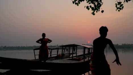 Una-Silueta-De-Bailarina-Masculina-Y-Femenina-Bailando-Danza-Clásica-India-Bharatnatyam-En-Un-Barco-Al-Atardecer-O-Al-Amanecer-En-La-Orilla-Del-Río-Ganga,-Cámara-Lenta