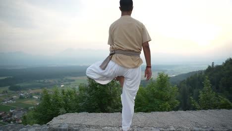 Indischer-Mann,-Der-Hatha-Yoga-Macht-Und-In-Der-Morgensonne-Bei-Sonnenaufgang-Am-Rand-Einer-Steinernen-Burgmauer-Steht-Und-Das-Tal-Mit-Feldern-Und-Wäldern-überblickt