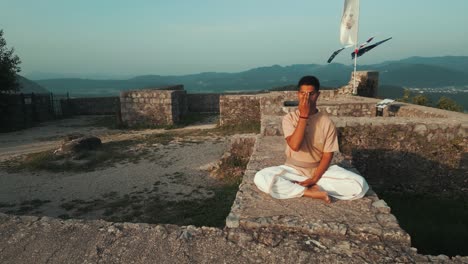 orbit-drone-shot-of-Indian-man-sitting-in-hatha-yoga-pose-and-meditating-nadhi-shuddhi-on-stone-castle-wall-on-top-of-the-hill-in-traditional-yogi-clothes-kurta-and-dhoti-at-sunrise