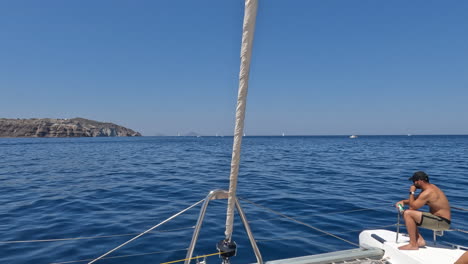 Onboard-a-yacht-sailing-past-the-greek-island-Thera-Santorini-whilst-a-man-sits-at-the-front-of-the-boat-on-a-beautiful-summers-day