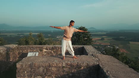 Indischer-Mann-Macht-Hatha-Yoga-Pose-Bei-Sonnenaufgang-Auf-Einer-Steinernen-Burgmauer-Auf-Dem-Gipfel-Des-Hügels,-Barfuß-Und-In-Traditioneller-Meditationskleidung,-Mit-Feldern-Und-Bäumen-Hinter-Ihm