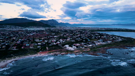 Sunset-drone-view-from-over-ocean-with-moody-clouds-of-Sandbaai,-Overstrand
