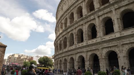 Looking-Up-At-Iconic-Colosseum-In-Rome-From-Piazza-Del-Colosseum-With-Tourists-Walking-Past-On-Sunny-Day-With-Blue-Skies-And-Light-Clouds