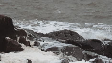 Slow-motion-waves-and-sea-foam-gather-around-well-worn-black-rocks-on-a-stormy-and-unsettled-day