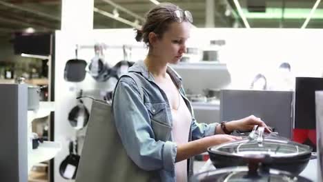 Handheld-Footage-of-a-young-woman-in-denim-shirt-shopping.-Looking-for-kitchen-stuff.-Examining-various-pots-and-pans-with-glass-top.-Supermarket,-store.-Side-view