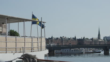 Swedish-flag-moves-in-wind-on-boat,-Stockholm-skyline-in-background