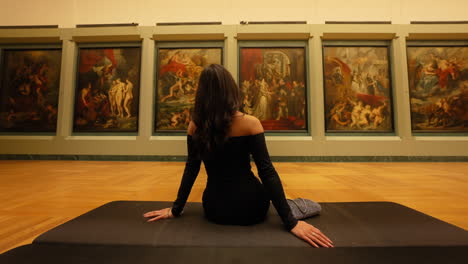 Back-View-Of-Female-Visitor-Sitting-And-Looking-At-Paintings-In-The-Gallery-Of-Louvre-Museum-In-Paris,-France
