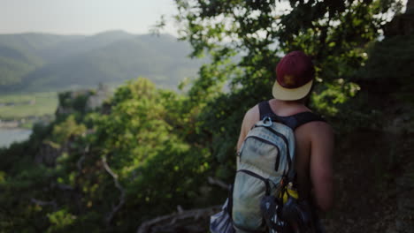 handsome-young-man-walks-mountain-landscape,-slow-motion,-male-with-backpack,-bare-chest,-hiking-trail,-mountain-route,-green-forest-valley,-sunny-weather,-austria,-durnstein,-europe,-cinematic-slomo