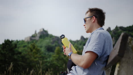 happy-smiling-man-drinking-bottle-of-water-from-a-thermos,-drinks-water,-Durnstein-castle-ruins-in-background,-mountain-valley-view,-Hydrating-male,-holiday-relaxation-trekking-hike-break,-friendly