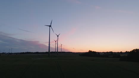 Aerial-establishing-view-wind-turbines-generating-renewable-energy-in-a-wind-farm,-evening-after-the-sunset-golden-hour,-countryside-landscape,-high-contrast-silhouettes,-drone-shot-moving-forward