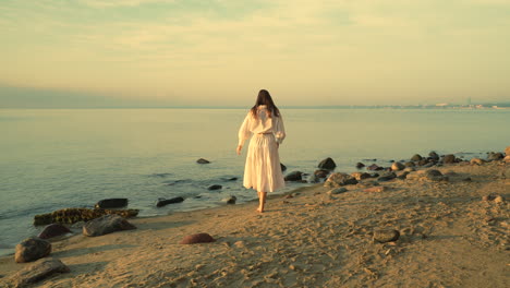 Caucasian-woman-wearing-a-beautiful-white-dress-walking-on-sand-beach-at-golden-sunset---Female-tourist-on-summer-vacation---Slow-motion-tracking