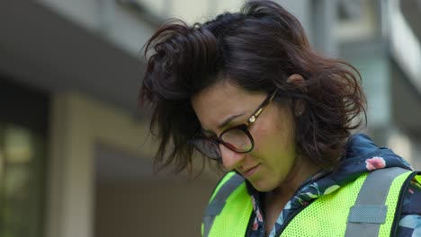 Young-brunette-woman-wearing-glasses-and-safety-vest-reads-focused-on-device-or-work-document