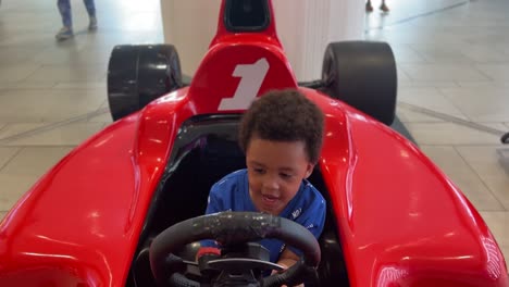 Fröhliche-Szene:-Ein-Dreijähriges,-Ausdrucksstarkes-Schwarzes-Kleinkind,-Das-In-Einem-Einkaufszentrum-Auf-Einem-Roten-Formel-1-Simulator-Fährt