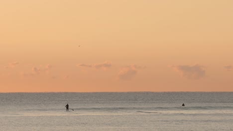 Paddle-Boarder-Und-Surfer-Im-Meer-Bei-Sonnenaufgang-Mit-Kleinen-Wellen
