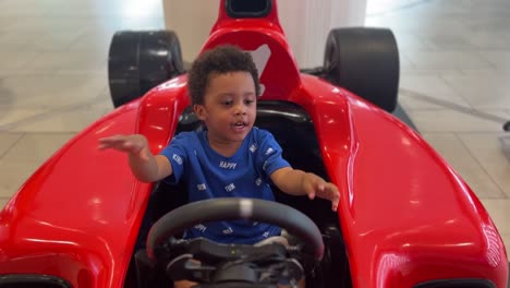 Joyful-scene:-3-year-old-very-expressive-black-child-enjoying-in-a-red-F1-simulator-inside-a-mall