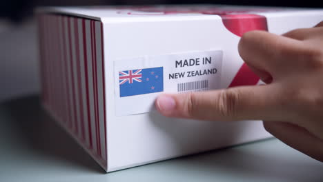 Manos-Aplicando-Etiqueta-De-Bandera-Hecha-En-Nueva-Zelanda-En-Una-Caja-De-Cartón-De-Envío-Con-Productos