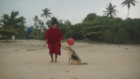 Indian-woman-stands-on-the-beach,-dressed-in-red-with-a-vibrant-balloon-in-hand,-she-playfully-interacts-with-the-wind-as-her-dress-dances-around-her