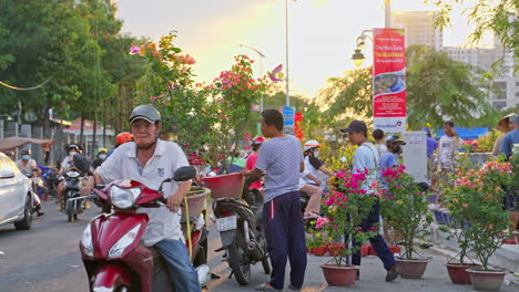 Vibrant-culture-of-Vietnam-as-locals-purchase-and-transport-paper-flower-pots-on-through-a-bustling-flower-market,-and-then-tie-them-on-their-motorbike-under-the-beautiful-evening-sunlight