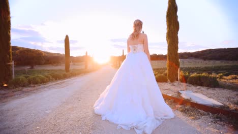 Bride-in-a-big-white-dress-standing-on-a-macadam-road-in-summer-at-sunset