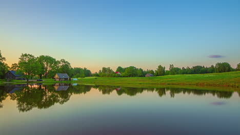 Timelapse-shot-of-sunset-in-the-background-along-village-cottages-with-landscape-reflecting-on-lake-surface-during-evening-time