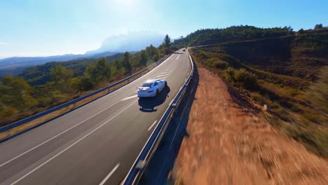 FPV-aerial-drone-following-a-white-Porsche-sportscar-driving-along-a-sunny-mountain-highway