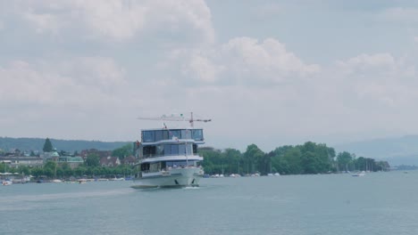 Scenic-drone-footage-of-a-ferry-boat-with-a-Swiss-flag-sailing-on-a-beautiful-lake-at-the-foot-of-the-Alps,-not-far-from-a-village-located-on-the-coast