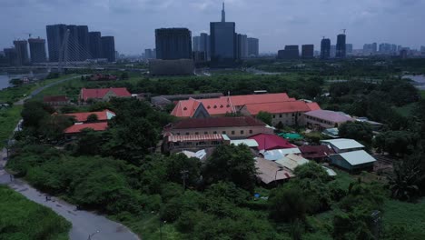 Thủ-Thiêm-Parish-Church-and-the-Lovers-of-the-Holy-Cross-Convent-are-the-oldest-French-Colonial-buildings-in-Ho-Chi-Minh-City,-Vietnam-Aerial-orbit-view-revealing-modern-skyline-in-the-background