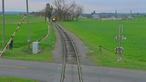 Una-Cabeza-En-La-Vista-De-Drones-De-Un-Antiguo-Tren-De-Pasajeros-De-Vapor-Que-Se-Acerca-Mientras-Viaja-A-Través-De-Tierras-De-Cultivo-En-Un-Día-De-Abril