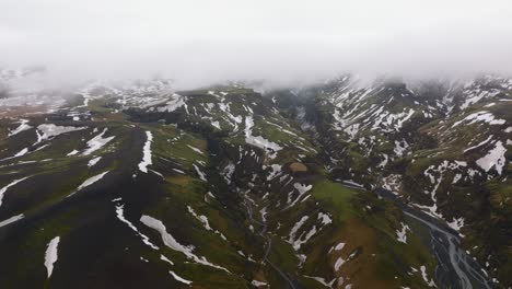 Aerial-view-of-rivers-flowing-through-mountains-covered-in-melting-snow,-on-a-cloudy-and-foggy-day,-in-Iceland