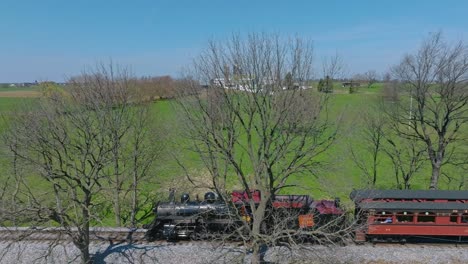 A-Drone-Side-View-of-a-Steam-Passenger-Train-Traveling-Blowing-Smoke,-on-a-Single-Track,-Traveling-in-Countryside-on-a-Sunny-Spring-Day