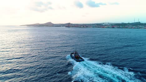 4k-Aerial-of-Tugboat-coming-into-Willemstad-city,-skyline-and-Queen-Juliana-bridge-in-background