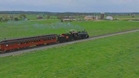 Ein-Drohnenblick-Von-Hinten-Auf-Einen-Dampfzug,-Der-An-Einem-Frühlingstag-In-Zeitlupe-Durch-Ackerland-Fährt