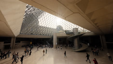 Wide-angle-shot-of-people-walking-under-the-famous-glass-pyramid-in-the-Louvre-Museum,-with-sunlight-coming-through-in-Paris,-France