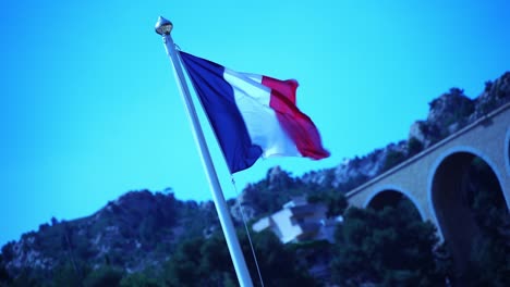 French-flag-waving-in-the-wind-in-front-of-a-beautiful-stone-railway-bridge-with-rocks-in-the-background