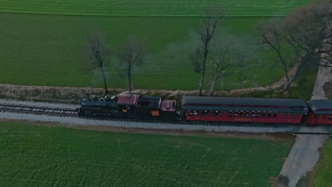 Una-Vista-Lateral-Aérea-De-Un-Tren-De-Pasajeros-De-Vapor-Que-Viaja-Soplando-Humo,-En-Una-Sola-Vía,-A-Través-De-Tierras-De-Cultivo-En-Un-Soleado-Día-De-Primavera