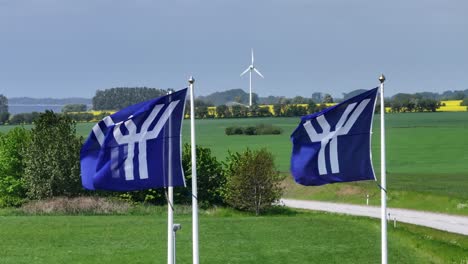 Static-closeup-blue-white-waving-Ystad-flags-with-panorama-view-and-wind-turbine