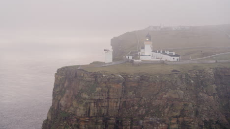 Aerial-shot-of-a-misty-lighthouse-glowing-at-dusk-atop-a-seaside-cliff