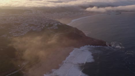 Wide-shot-of-Nazaré-town-Portugal-during-sunrise-with-low-clouds,-aerial