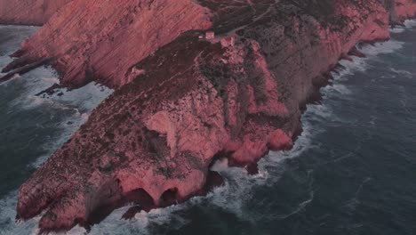 Reveal-shot-of-high-cliff-at-Farol-do-cabo-Espichel-Portugal-with-sunrise,-aerial