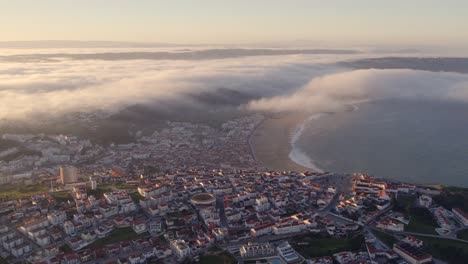 Low-clouds-rolling-over-coastline-at-Nazare-Portugal-during-sunrise,-aerial