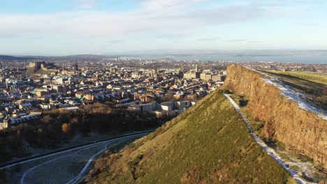 Aerial-shot-of-Edinburgh-in-the-snow,-with-Arthurs's-seat-and-Salisbury-crags-in-the-foreground,-and-Edinburgh-Castle-in-the-background